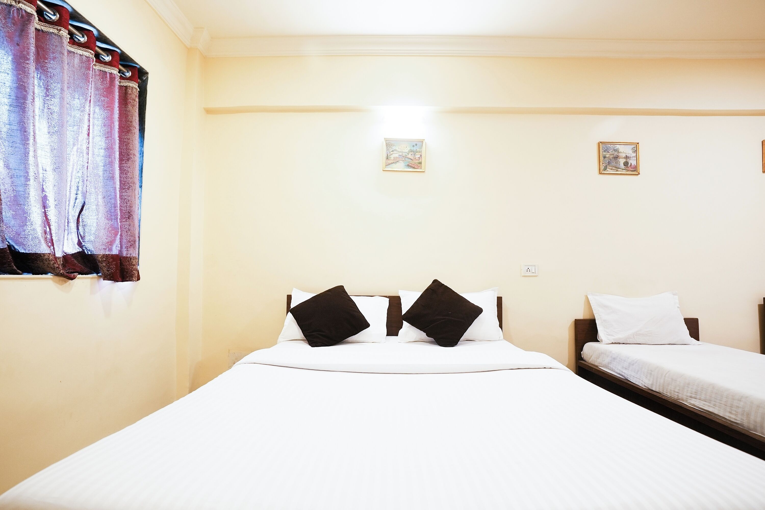Habitación de hotel con una cama grande, dos camas más pequeñas, una ventana con cortinas y dos cuadros colgados en la pared.