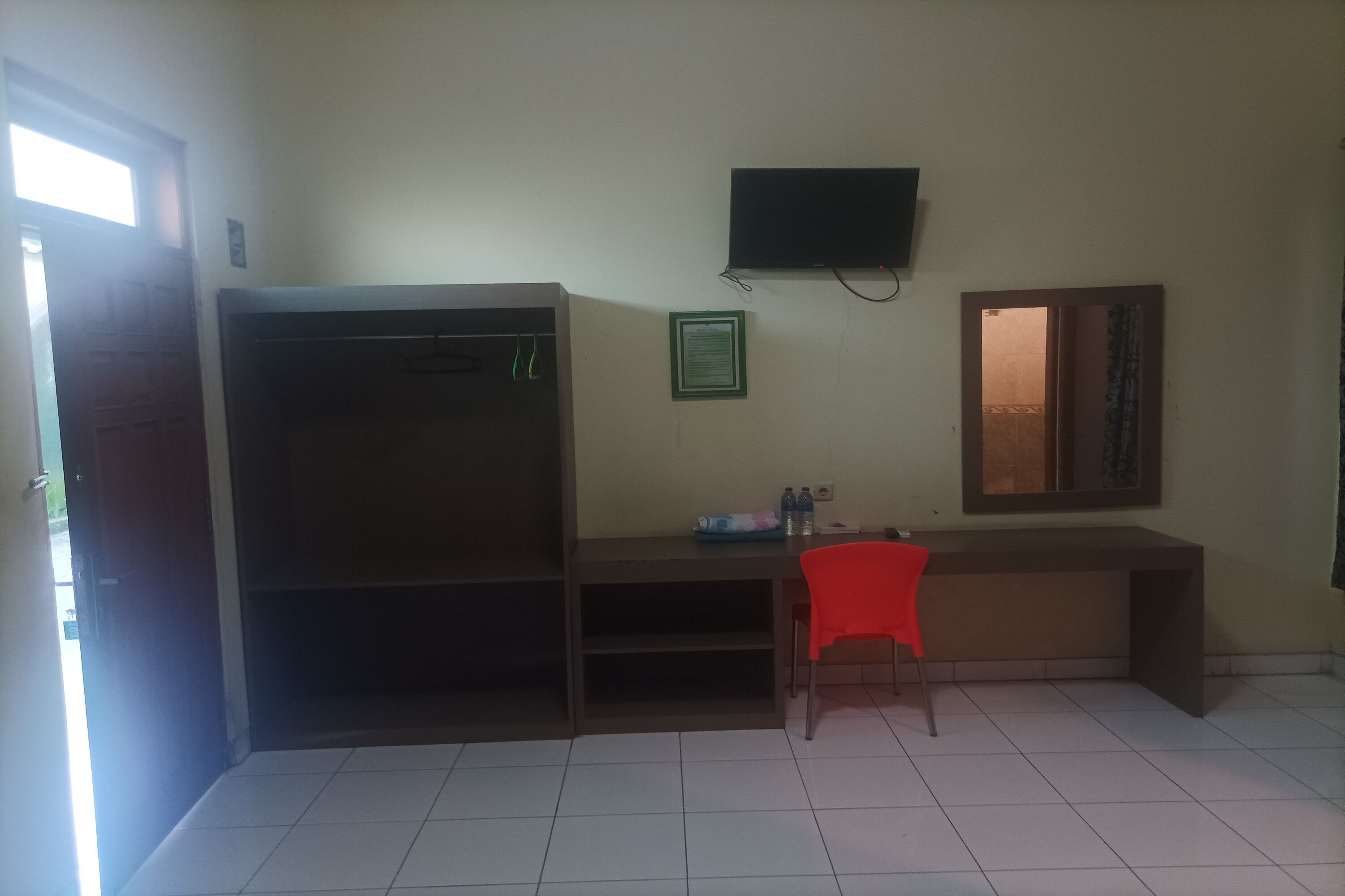 Una habitación con escritorio, una silla roja, un armario, un televisor montado en la pared y una puerta que da al exterior.