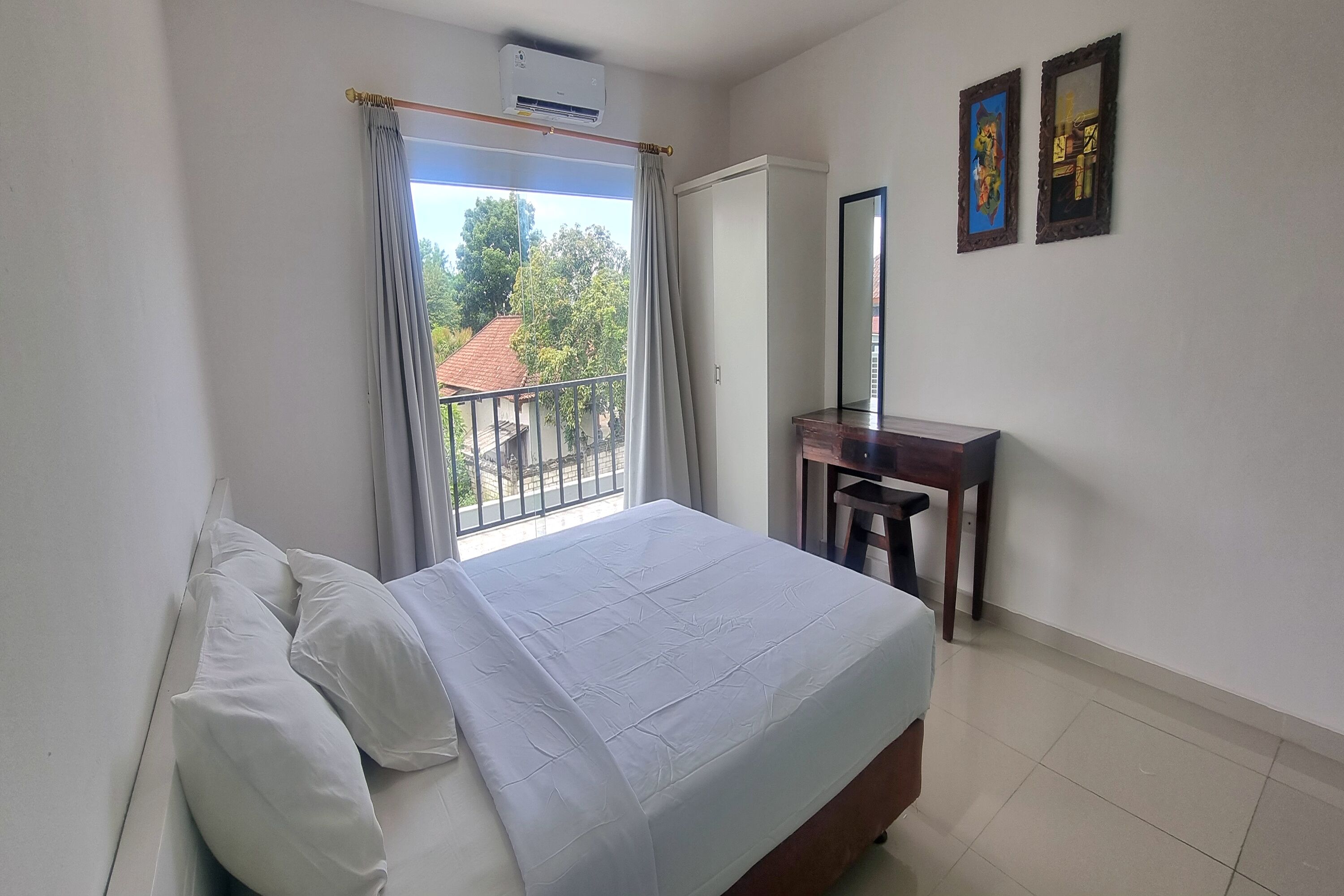 A bedroom with a bed, a desk, a mirror, and a view of the outside.