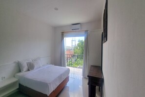 A hotel room with a bed, a small table, and a balcony with a view of buildings and greenery. - Tresna Villa 7 (Nusa Dua)