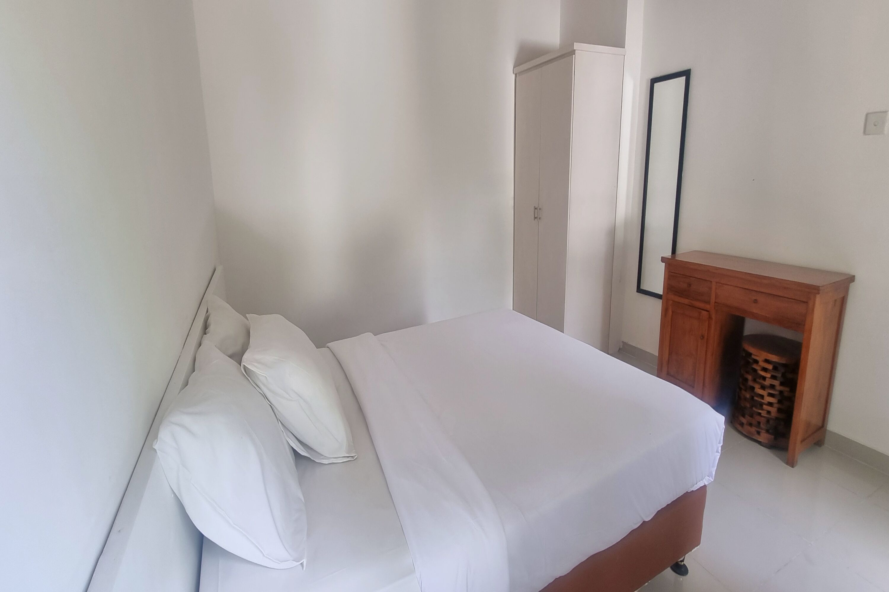 Ein ordentlich bezogenes Bett mit weißen Leinen in einem Zimmer mit weißen Wänden, einem Holfnachtschränkchen und einem Kleiderschrank.