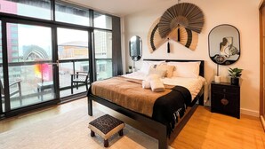A modern bedroom with a large window, a bed with white linens, a small stool, and a decorative fan. - Hydro Apartments (Glasgow)