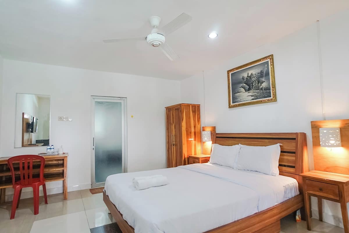 A hotel room with a bed, a desk with a chair, a wardrobe, a ceiling fan, and a framed picture on the wall.