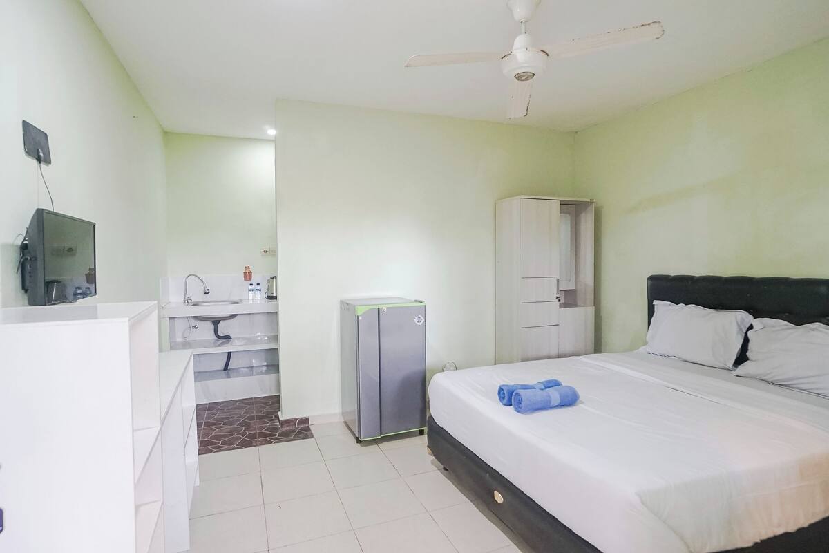 A hotel room with a bed, a desk, a wardrobe, a small refrigerator, and a ceiling fan.