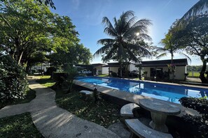 Piscina com um caminho de pedra que leva a um edifício, cercada por vegetação exuberante e palmeiras.