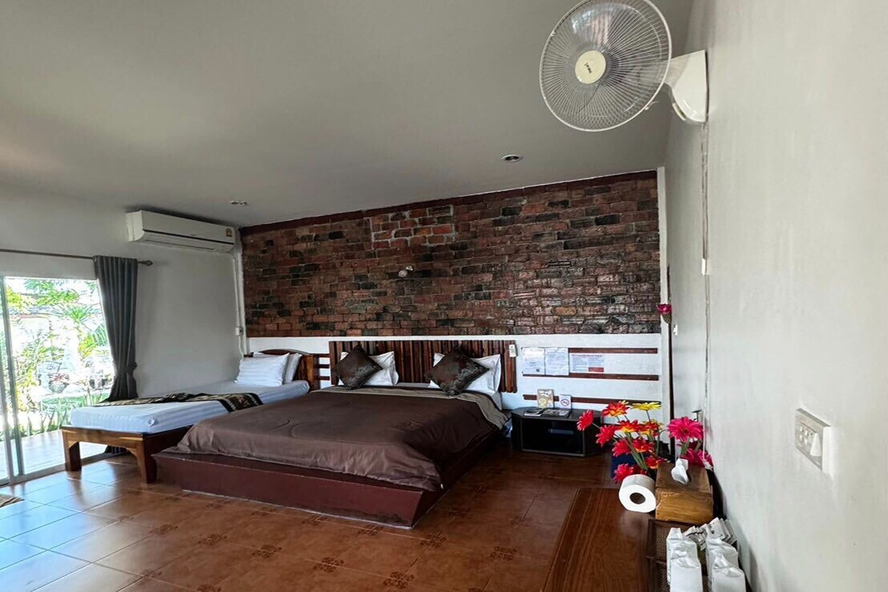 Habitación de hotel con pared de ladrillo, una cama grande, otra más pequeña, una mesita de noche con lámpara, un ventilador y vistas a zonas verdes.