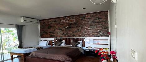 Habitación de hotel con pared de ladrillo, una cama grande, otra más pequeña, una mesita de noche con lámpara, un ventilador y vistas a zonas verdes.