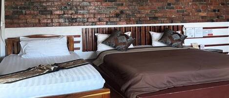 A room with two beds, one with a wooden frame and the other with a metal frame, both with brown bedding. A brick wall is behind the beds, and there is a wooden headboard.