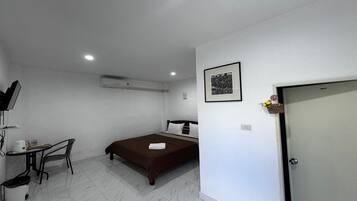 A small, clean room with a bed, a desk, and a chair. There is a picture on the wall and an air conditioning unit above the bed.