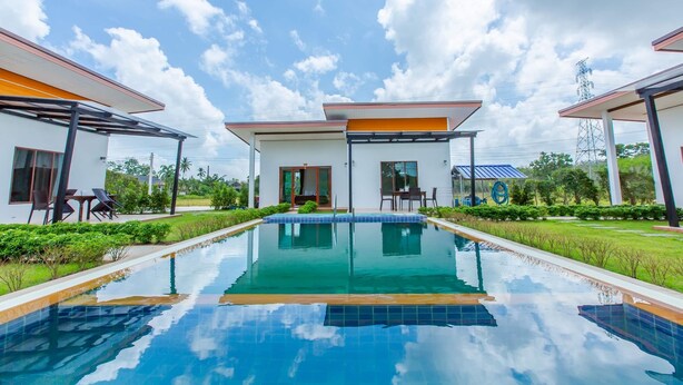 Un área de piscina moderna con agua azul cristalina, rodeada de vegetación cuidada y edificios contemporáneos con grandes ventanales y mobiliario exterior.