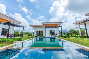 Un área de piscina moderna con agua azul cristalina, rodeada de vegetación cuidada y edificios contemporáneos con grandes ventanales y mobiliario exterior.
