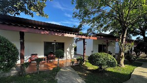 Un edificio de dos pisos con techo de tejas, un patio cubierto con mesa y sillas de madera, y un jardín cuidado con arbustos variados y un sendero.