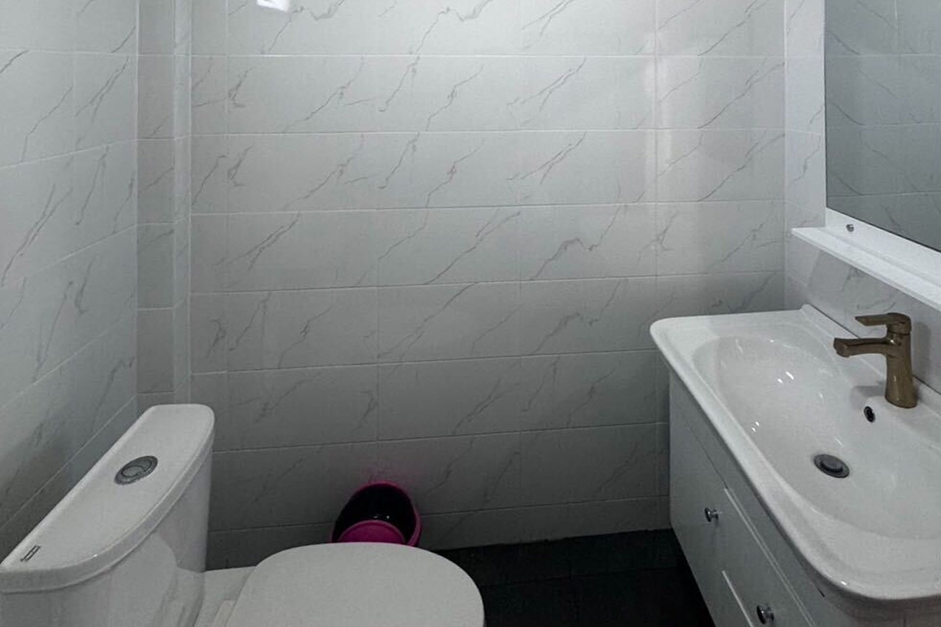 Un baño con baldosas de mármol blanco, inodoro blanco, lavabo blanco con grifo dorado y un espejo sobre el lavabo.