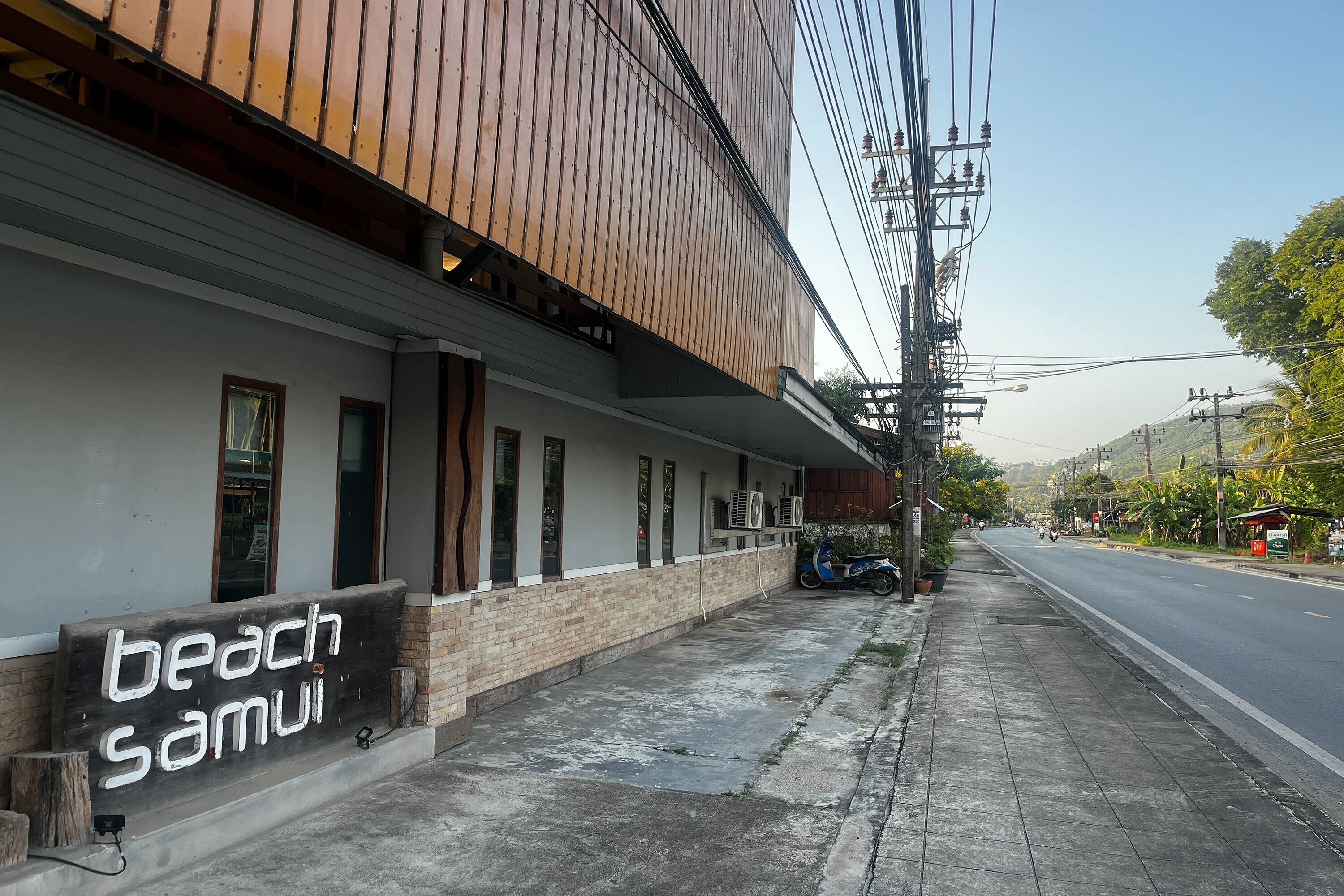 A building with a sign reading "beach samui" and a sidewalk with a parked scooter.