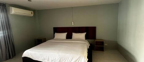 A hotel room with a bed, a nightstand, an air conditioning unit, and a wall-mounted light fixture.