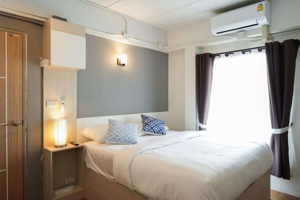 A hotel room with a bed, bedside table, lamp, and an air conditioning unit.