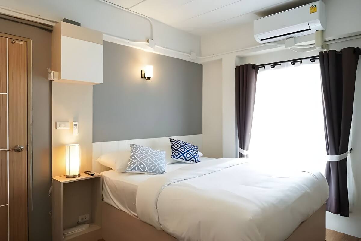 A hotel room with a bed, bedside lamp, air conditioning unit, and a window with curtains.