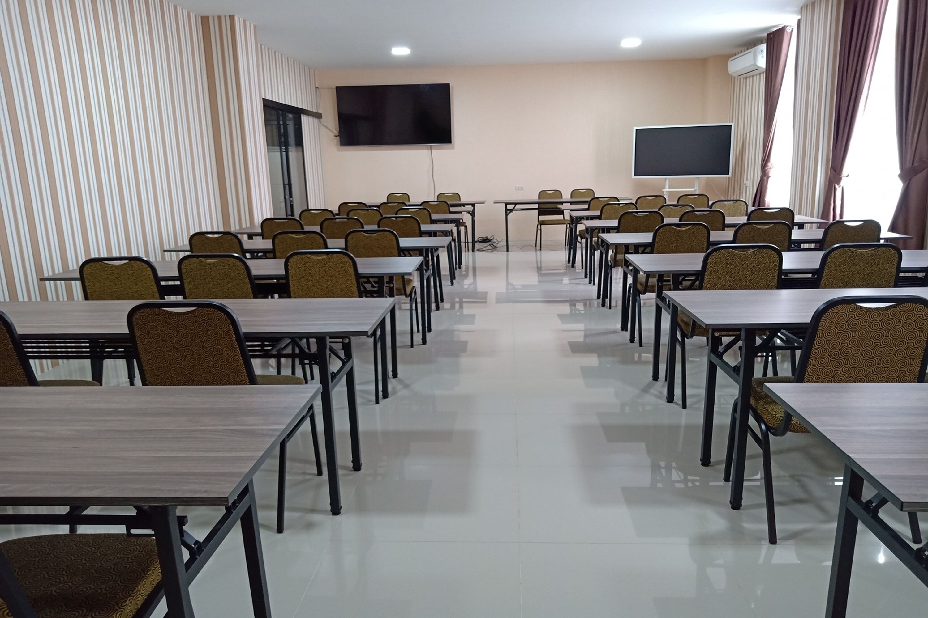 A conference room with rows of tables and chairs, a large flat-screen TV, and a window with curtains.