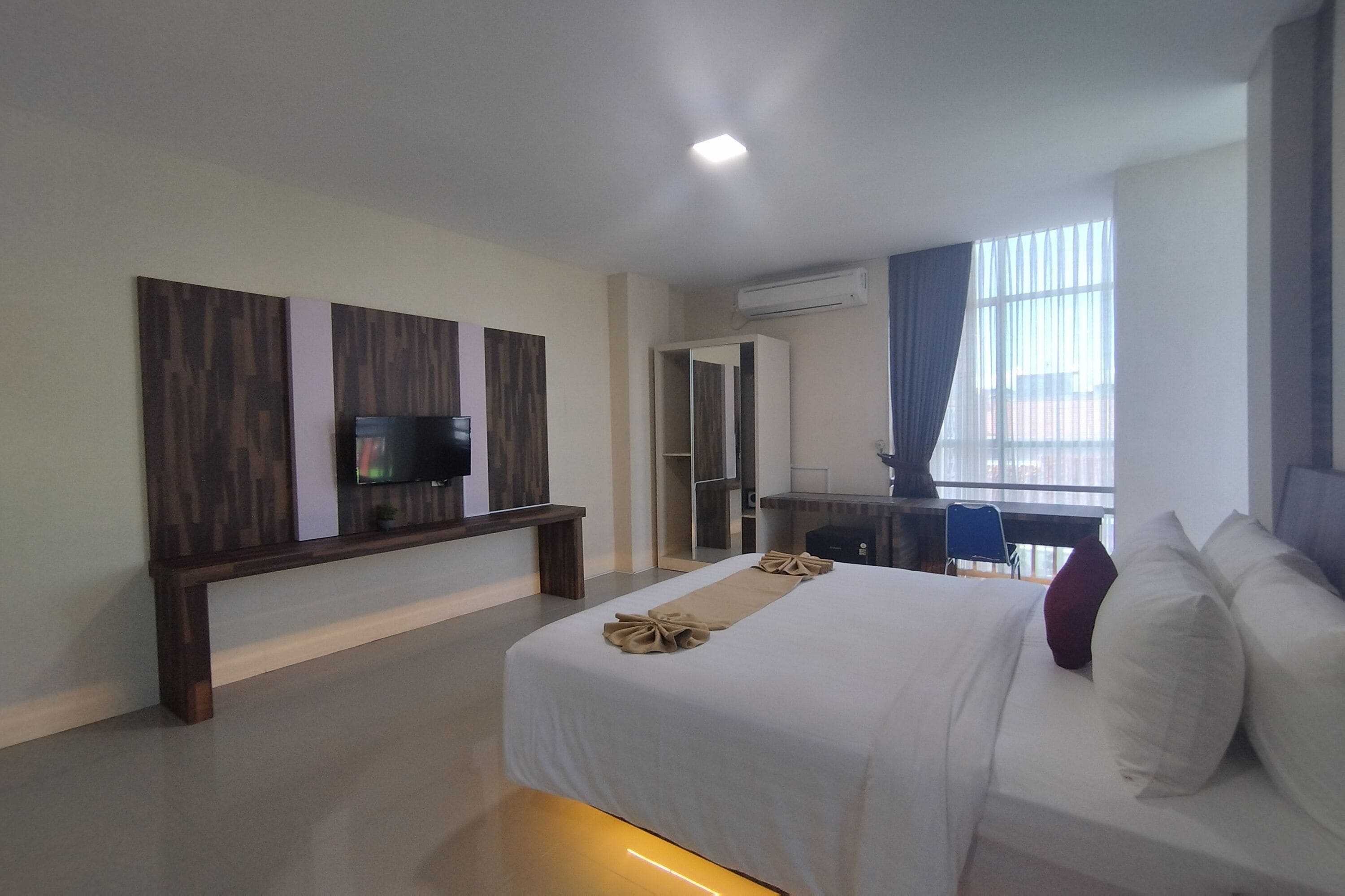 A modern hotel room with a large bed, a TV mounted on the wall, a desk, and a window with curtains.