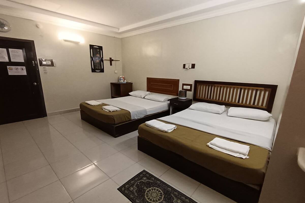Quarto de hotel com duas camas, uma mesinha de cabeceira, uma luminária fixada na parede e uma porta.