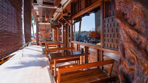 Un espacio al aire libre con bancos y mesas de madera, un muro de madera con un diseño tallado y vista a la calle.