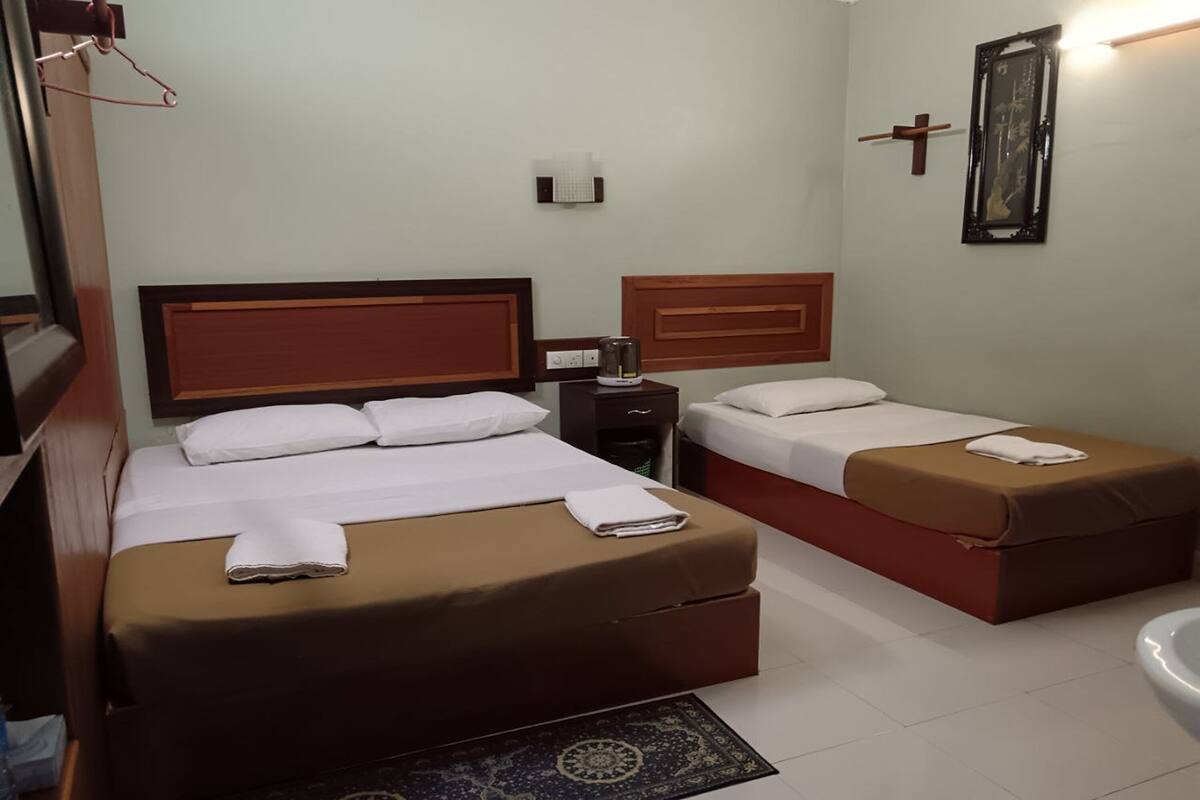 Quarto de hotel com duas camas, uma mesa de cabeceira, uma mesinha, uma luminária na parede, uma cruz e um quadro emoldurado.