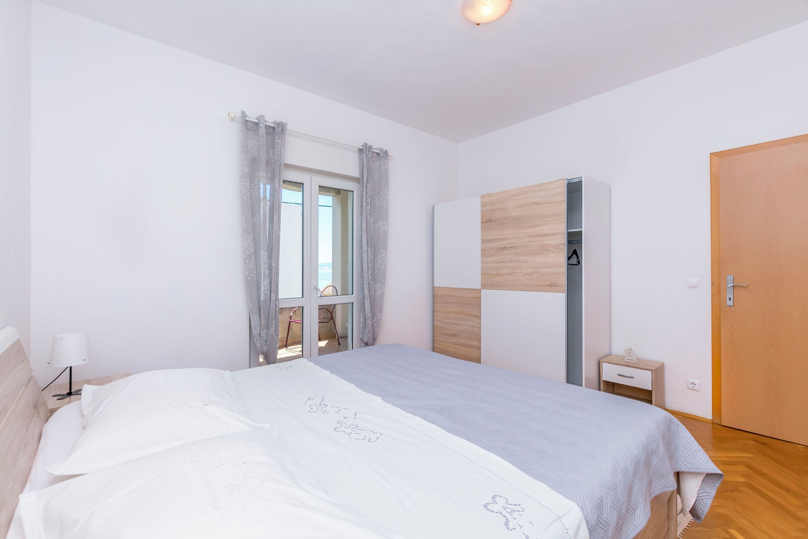 A bedroom with a bed, a window with curtains, a wardrobe, a bedside table, and a door.
