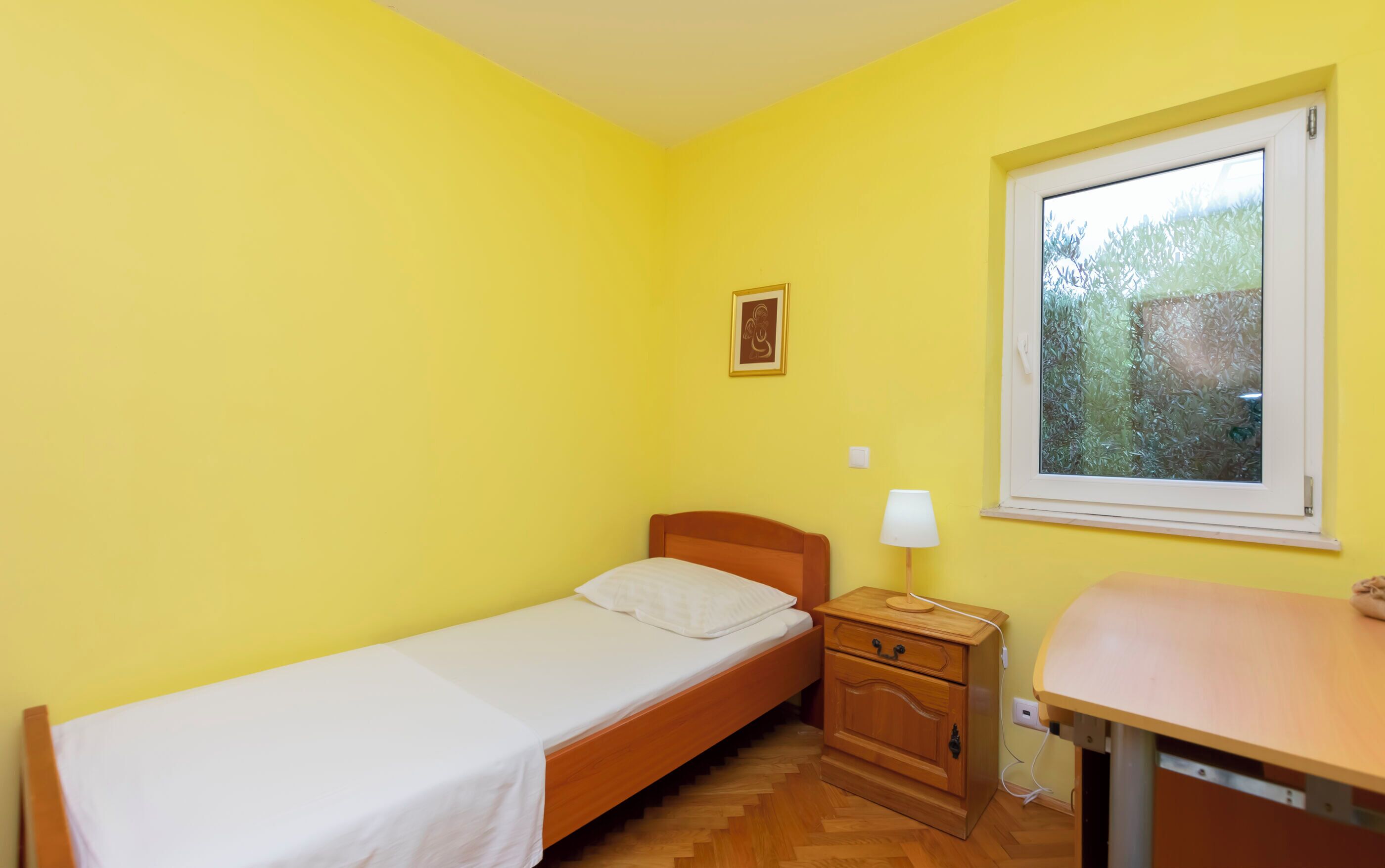 A small, single-bed room with a wooden bed, a bedside table with a lamp, and a window with a view of trees.