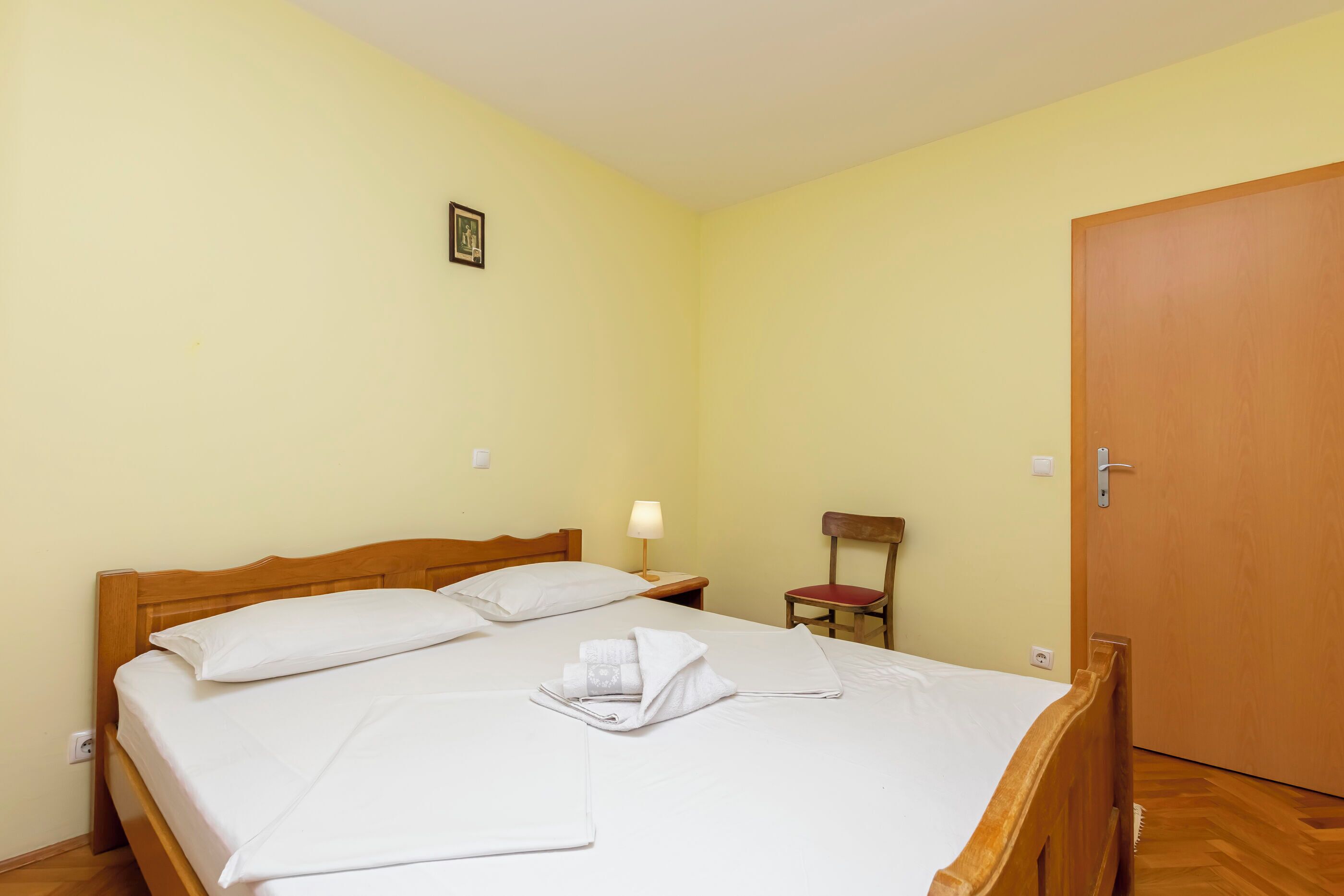 Una cama bien hecha con sábanas y toallas blancas, un cabecero de madera, una mesita de noche con una lámpara, una silla y una puerta.