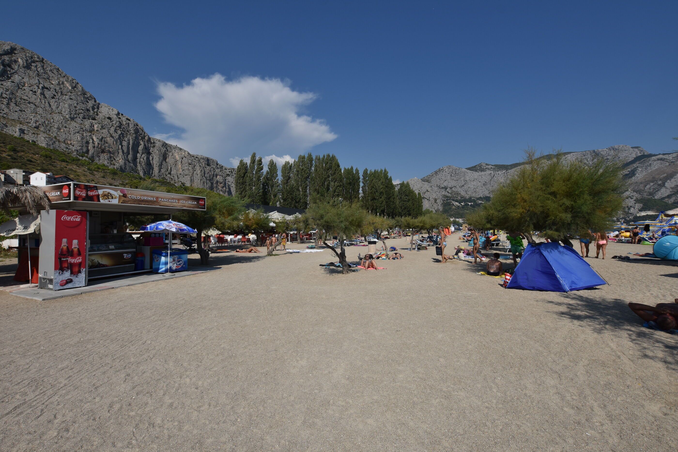 A beach with a Coca-Cola kiosk, a blue tent, and people relaxing.