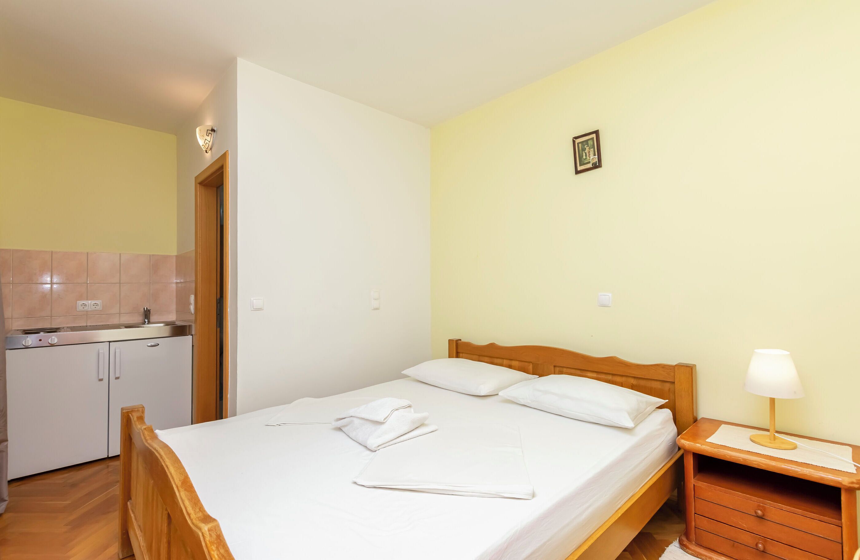 A clean, well-lit room with a wooden bed, white linens, a bedside table with a lamp, and a small kitchen area with a sink and cabinets.