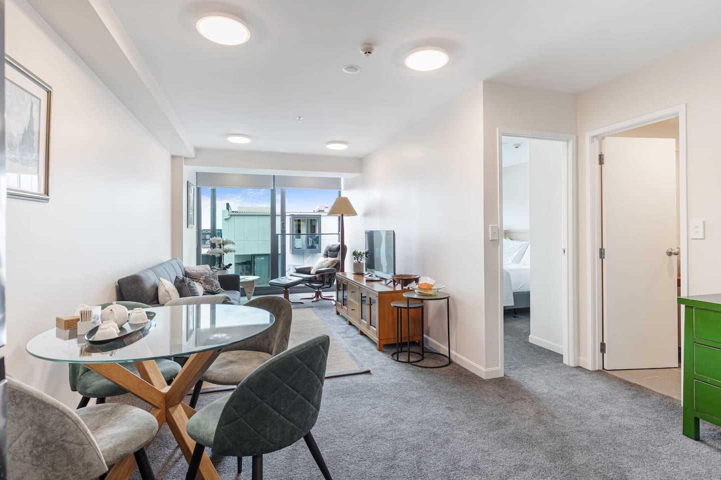 A modern hotel room with a dining area, a sofa, a small kitchen, and a view of the outside.