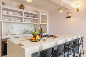 A kitchen with white shelves, a tiled backsplash, a fruit bowl, and bar stools. - Apartments Sonata (Omis)