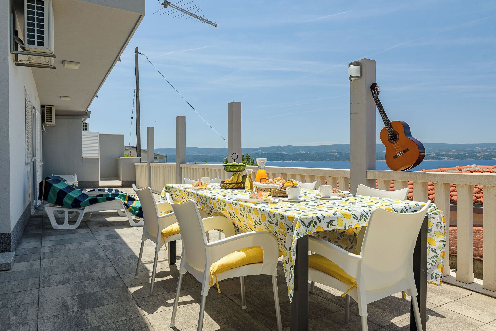Terrassa amb taula preparada per esmorzar, cadires, una guitarra i vista al mar.