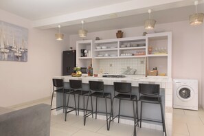 A modern kitchen with a central island, bar stools, a washing machine, and a dining area with a grey couch. - Apartments Sonata (Stanici)