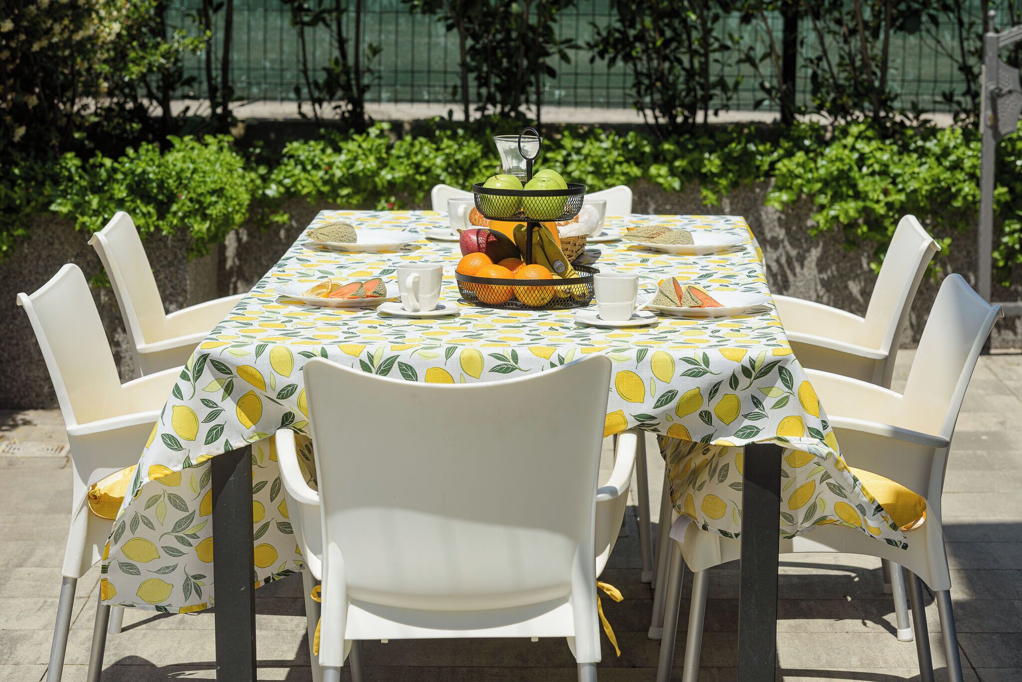 A table set outdoors with a fruit basket, white chairs, and a tablecloth with a lemon pattern.