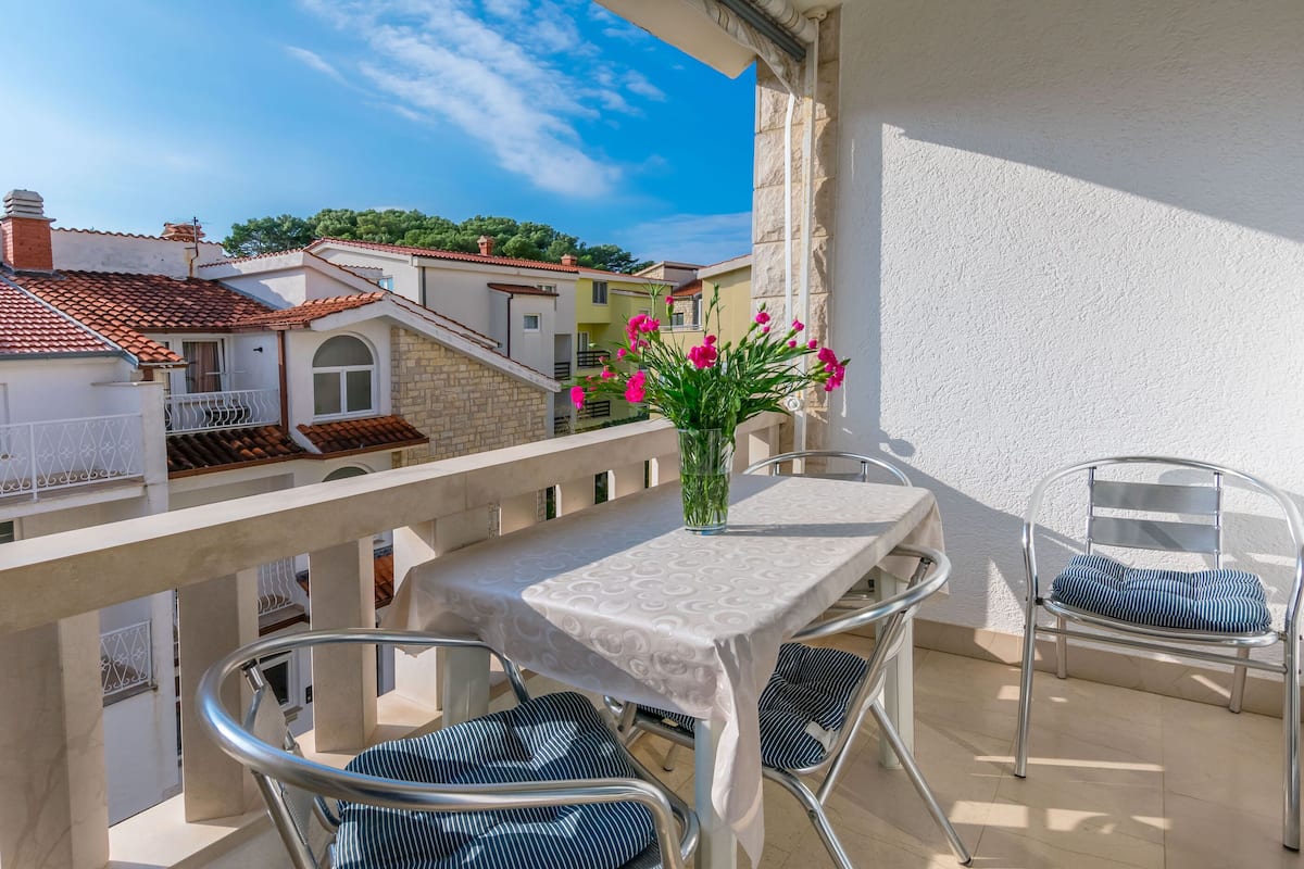 A balcony with a table and chairs, a vase with flowers, and a view of neighboring buildings.
