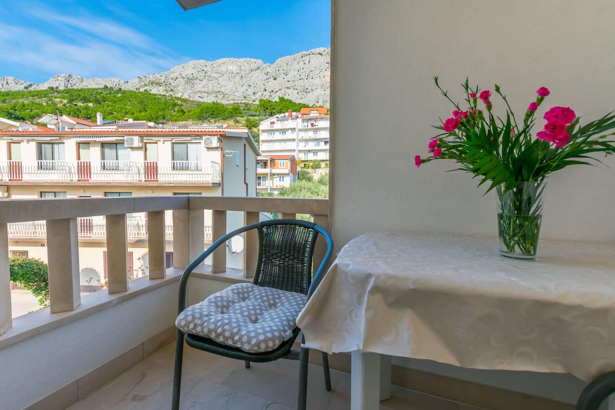 A balcony with a chair and a table, a vase with flowers, and a view of buildings and mountains.