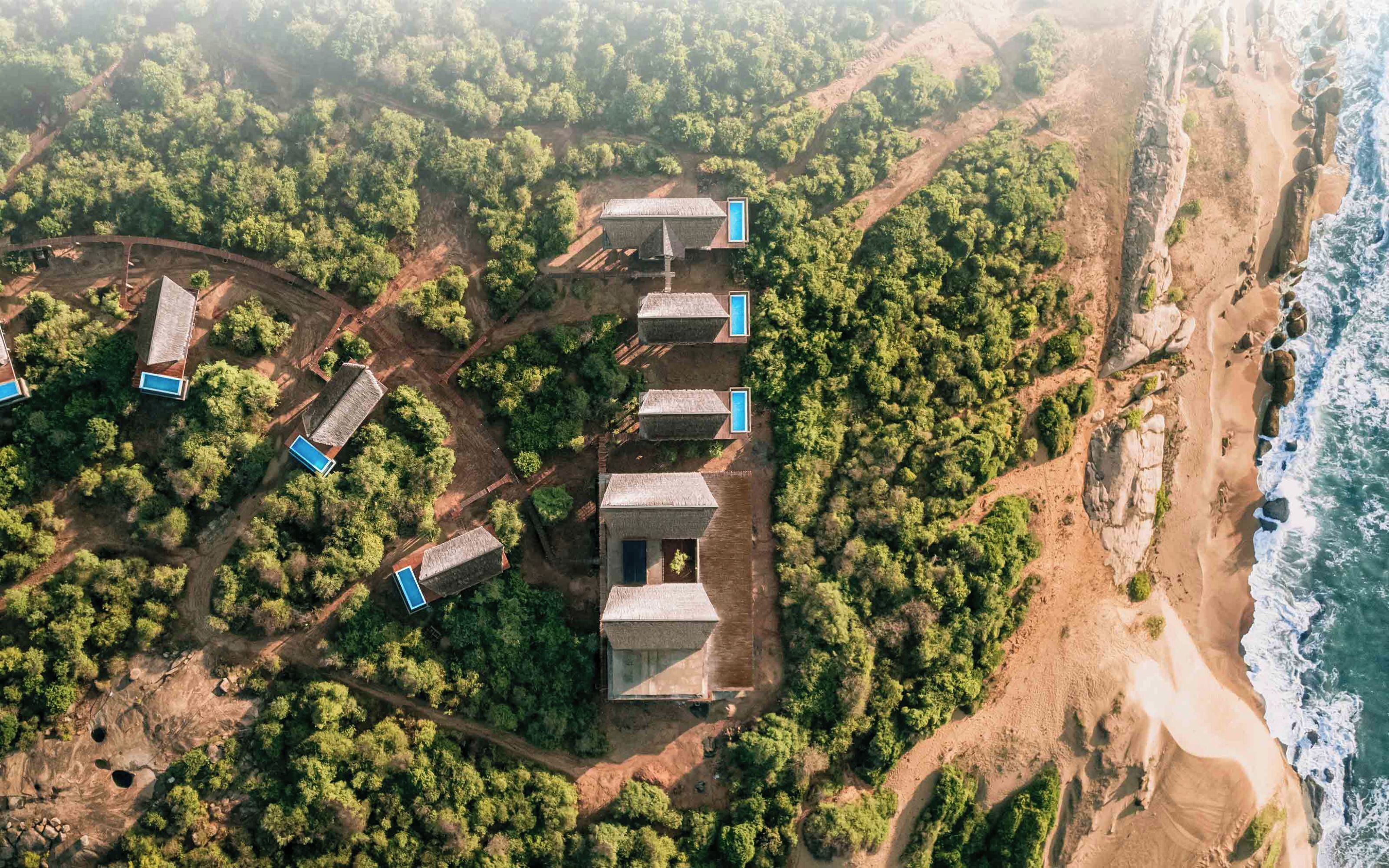 Une vue aérienne d’une station balnéaire dotée de plusieurs bungalows aux toits de chaume, chacun ayant sa propre piscine privée, le tout entouré d’une végétation luxuriante et bordé d’une côte rocheuse.
