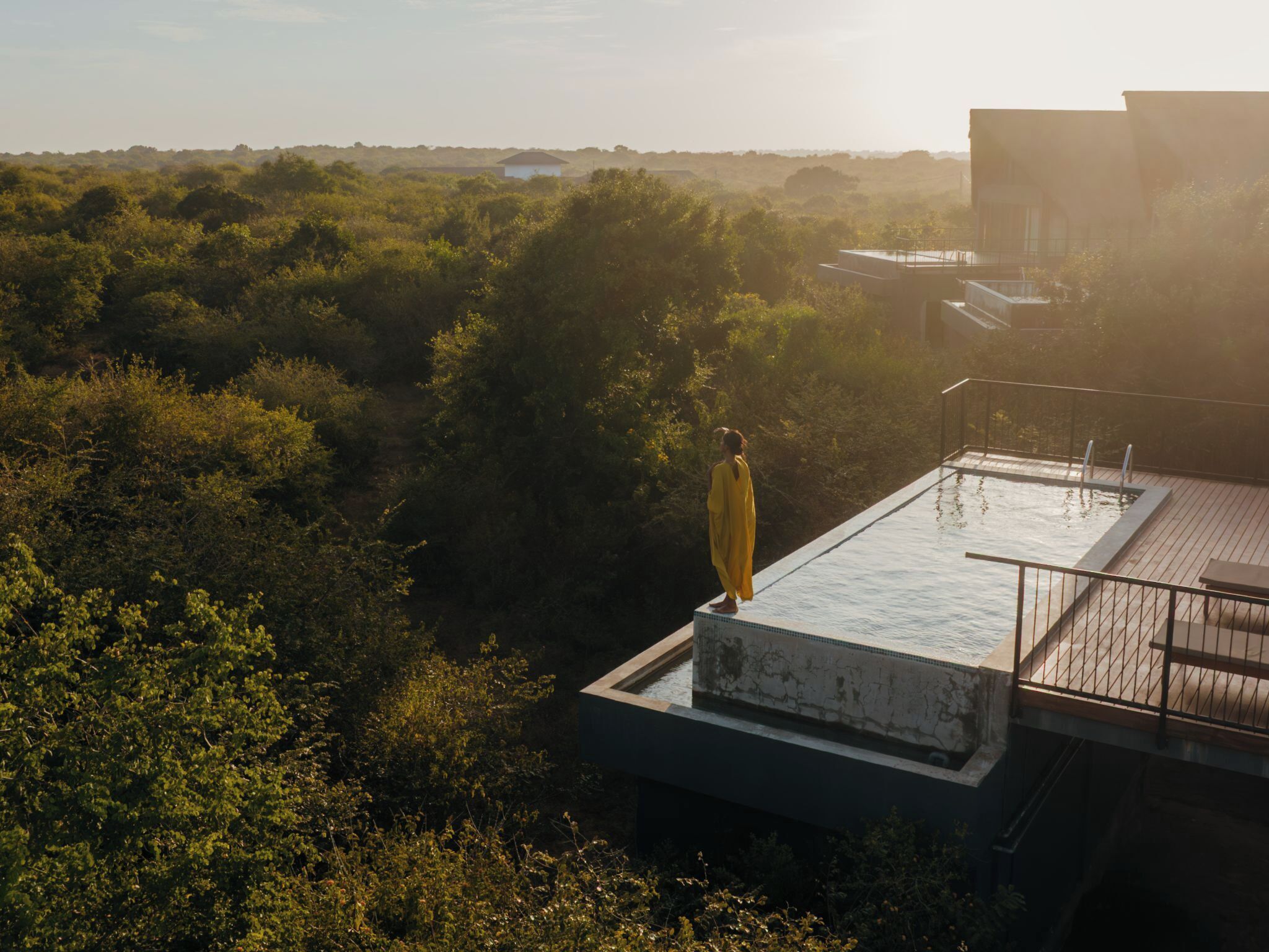 A person in a yellow robe standing on a rooftop pool with a view of dense greenery and distant buildings.