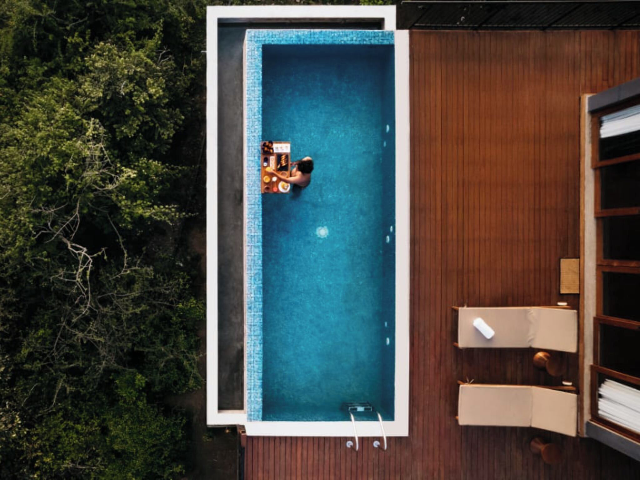 Eine Vogelperspektive zeigt einen rechteckigen Pool mit einer umlaufenden Holzterrasse, einem kleinen Tisch mit Stühlen und einer Holzfassade mit einem Fenster.