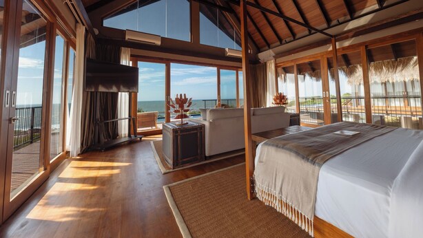 A spacious bedroom with a large bed, a sitting area with a table, and a view of the ocean through large windows. - Kotiyagala Luxury Villas (Palatupana)
