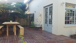 Una terraza de madera con una mesa y bancos, una puerta blanca con celosía decorativa y un ventanal de hojas múltiples.