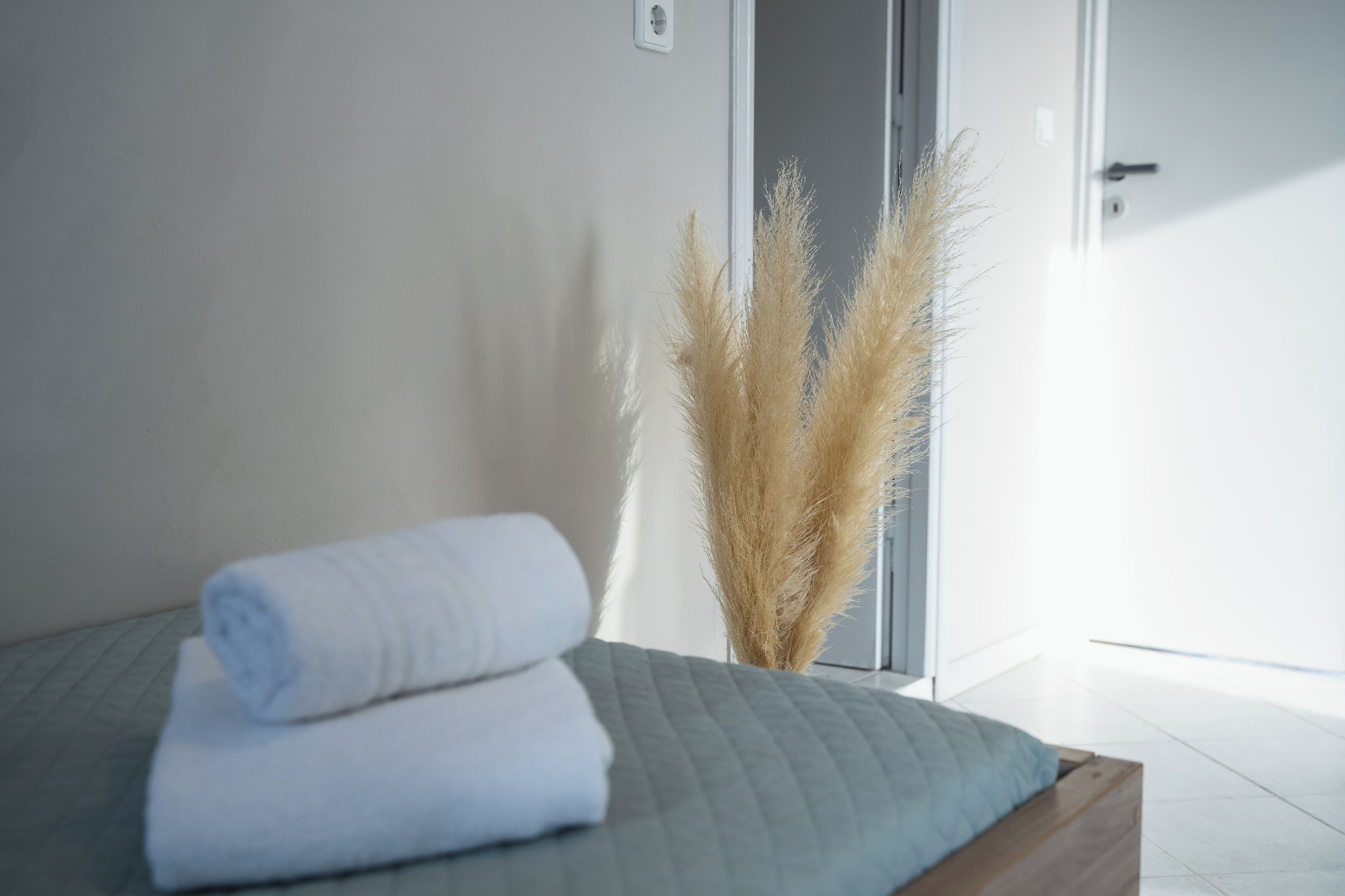 A neatly made bed with rolled white towels, a green quilted bedspread, and pampas grass in a vase.