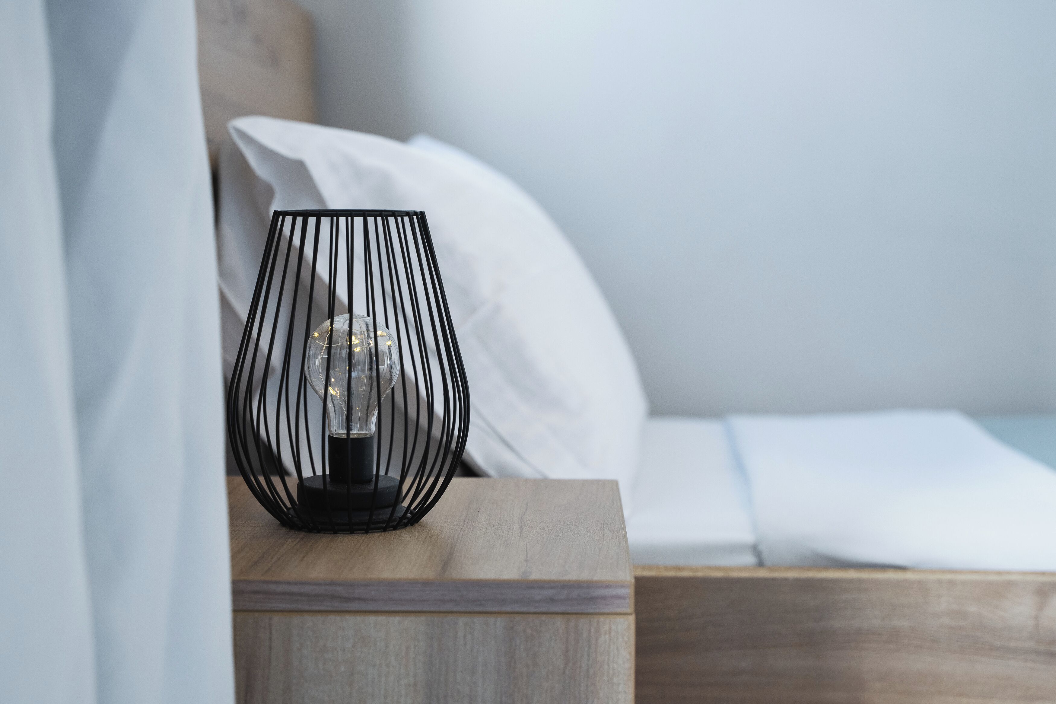 A bedside table with a black wireframe lamp and a white pillow on a wooden bed.