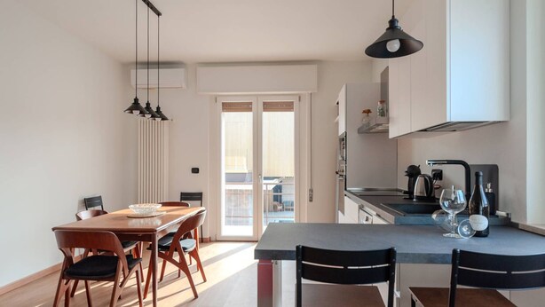 A modern kitchen with a dining area, featuring a large island, pendant lights, and a view to an outdoor balcony.