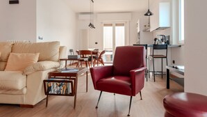 A modern living room with a beige sofa, a red armchair, and a wooden coffee table. There is a dining area with a table and chairs, and a kitchen area with a sink and countertop.