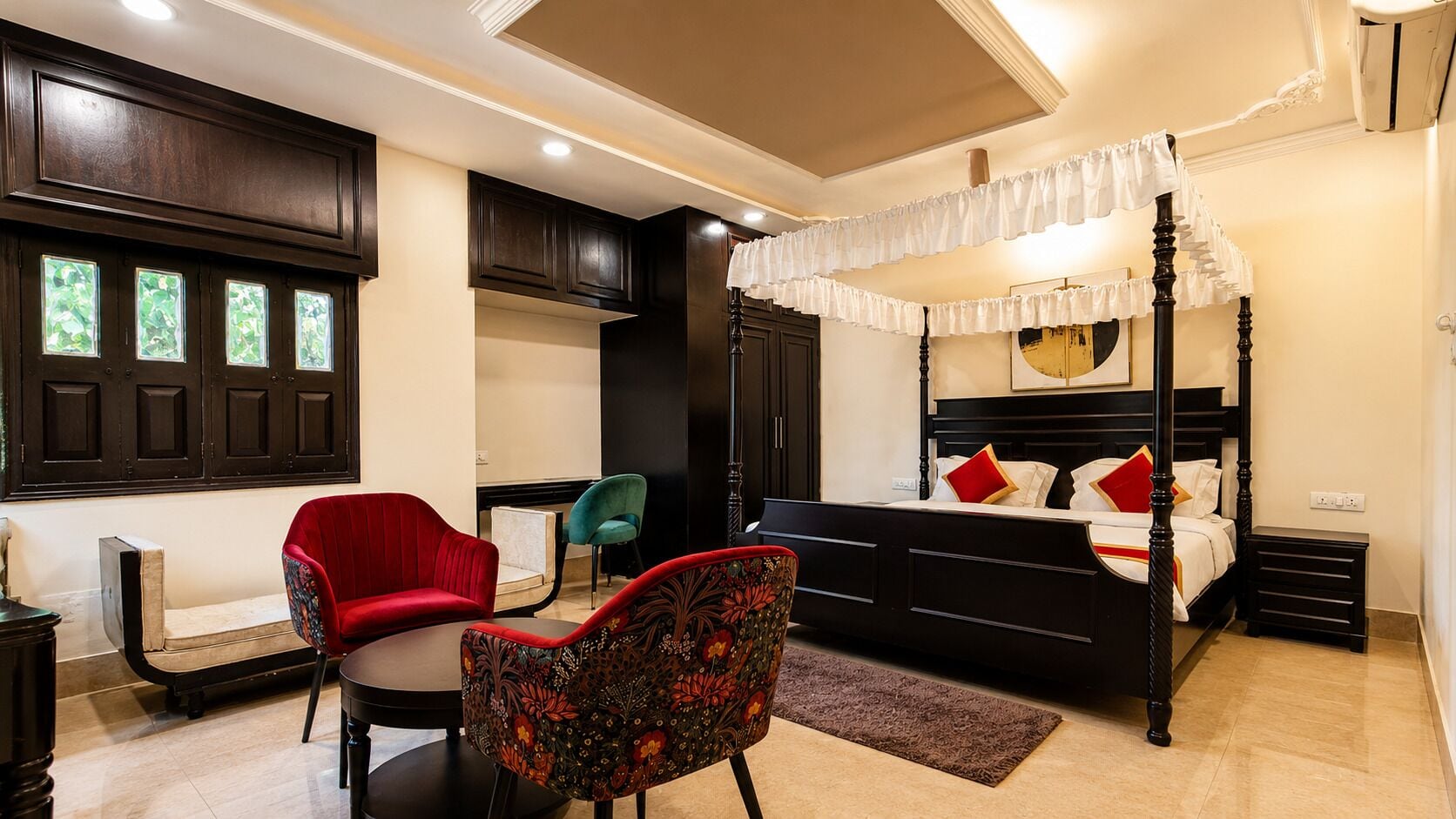 A bedroom with a four-poster bed, a red chair, a teal chair, and a small table.