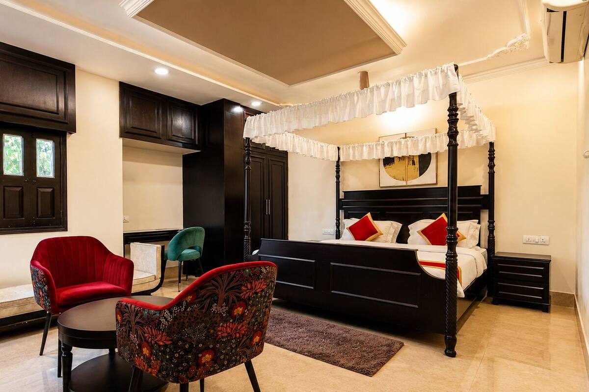 A bedroom with a four-poster bed, a red chair, a teal chair, and a small table.