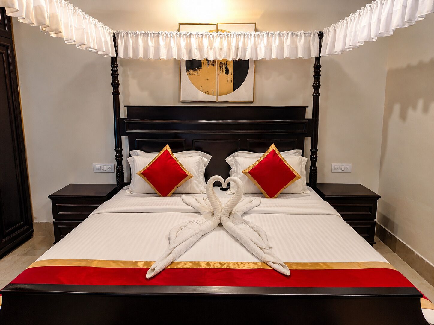 Una habitación de hotel con una cama con dosel, dos mesitas de noche, un corredor de cama rojo y dorado, y un cuadro colgado en la pared.